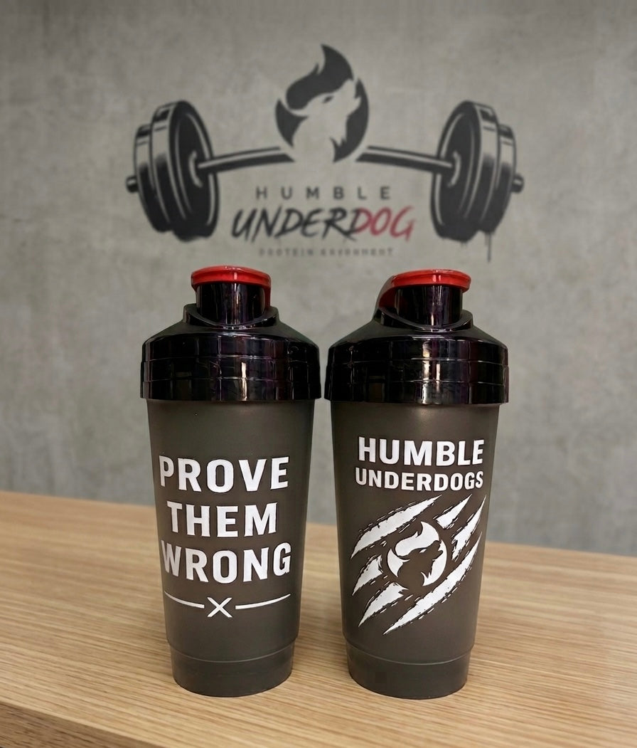 Two black shaker bottles with red caps displayed on a wooden surface against a gray wall with a 'HUMBLE UNDERDOG' logo. The bottle on the left reads 'PROVE THEM WRONG' and the bottle on the right reads 'HUMBLE UNDERDOGS' with a claw graphic.